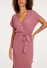 Vestido wrap en tela texturada de color malva con escote en V profundo y mangas cortas. Presenta un cinteo en la cintura y detalle de bolsillo lateral.