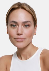 Gros plan sur une personne portant de petites boucles d'oreilles en or. Elle a la peau claire, une texture lisse et porte un haut sans manches blanc.