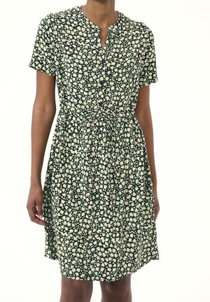 Kobieta nosząca ciemnozieloną, krótką sukienkę do kolan z małym wzorem białych stokrotek i zapinana na guziki z przodu.