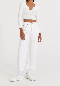 Kobieta w białej, długiej bluzce typu crop, białych, wysokich spodniach o szerokich nogawkach, białych skarpetkach i białych trampkach na jednolitym tle.