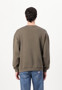 Sweatshirt vert olive avec une coupe décontractée, col et poignets côtelés, et fentes latérales. Assorti avec un jean légèrement déchiré bleu clair.