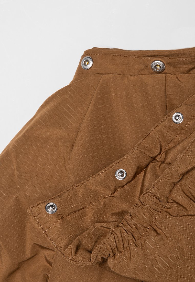 Brauner Stoff mit gestreiftem Muster, mit einer Rüsche am Rand und silbernen Druckknöpfen zum Schließen.