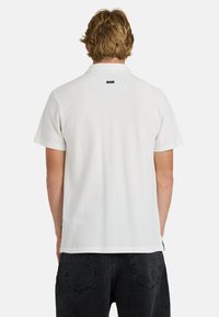 Άνδρας με κοντά ξανθά μαλλιά, φοράει απλό λευκό πόλο με κοντά μανίκια και μαύρα τζιν, φωτογραφημένος από πίσω σε λευκό φόντο.