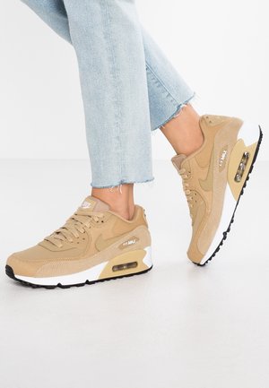 Jean bleu clair avec ourlets effilochés et baskets Nike Air Max couleur fauve avec semelles intermédiaires blanches et semelles extérieures noires sur un sol blanc.