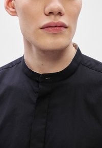 Fekete, magas nyakú ing, puha anyagból készült, elülső záródással és minimalista dizájnnal, sima textúrát és modern sziluettet bemutatva.