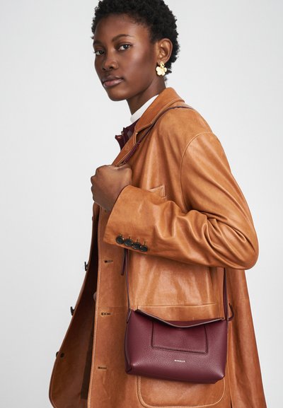 Bolsa de couro bordô com um design minimalista, apresentando uma aba frontal e uma alça de ombro, acompanhada de um casaco de couro castanho.