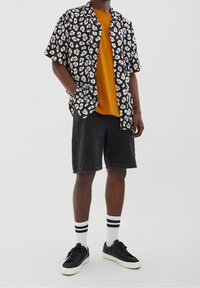 Floral-printed short-sleeve shirt in black, orange, and white, layered over a mustard yellow t-shirt, paired with black denim shorts and black sneakers.