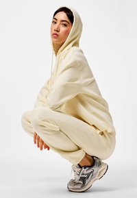 Mlada ženska v počepu, oblečena v kremasto kapucar in ujemajoče se športne hlače, z sivimi in črnimi tekaškimi copati na belem ozadju.