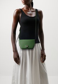 Zelená kožená kabelka přes rameno s geometrickým tvarem a stříbrným kulatým uzávěrem. Nesená přes jednobarevný černý top a bílou sukni.