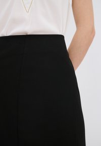 Musta hame, jossa on sileä kangas, korkea vyötärö ja istuva malli. Yhdistettynä valkoiseen toppiin, jossa on V-pääntie ja pehmeä kangas.