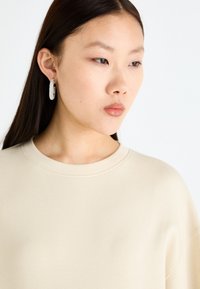 Beige genser med rund hals, myk tekstur og avslappet passform. Modellen har på seg sølvfargede hoop øredobber pyntet med små klare krystaller.