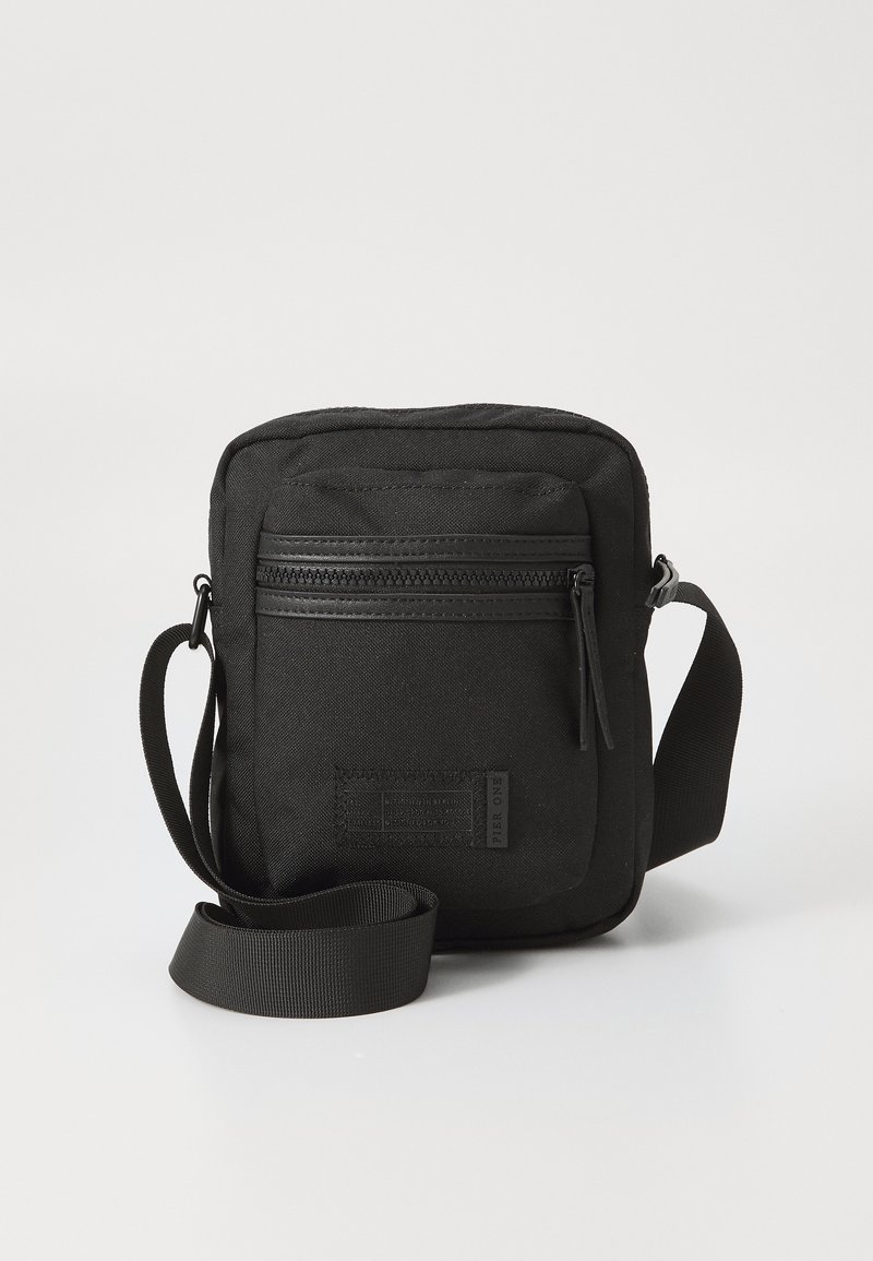 Borsa a tracolla nera realizzata in tessuto testurizzato, con una tasca anteriore con zip e una tracolla regolabile. Presenta un piccolo patch con il logo sulla parte anteriore.