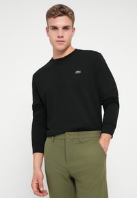 Crna dugih rukava majica s okruglim izrezom, s malim zelenim Lacoste logotipom. Uparena s maslinastozelenim hlačama, glatke teksture.