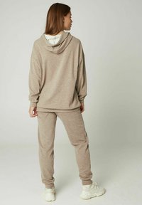 Beige sweatshirt en bijpassende broekset, met een capuchon, lange mouwen en geribde manchetten, gecombineerd met witte sportschoenen.