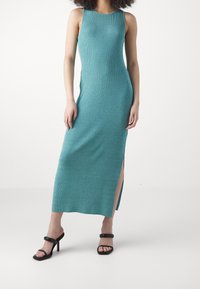 Abito maxi a coste color turchese con scollatura a barca, spacco laterale e silhouette aderente. Abbinato a sandali neri con tacchi a strappo.