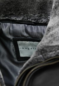 Sivá kabátová podšívka s jemným, štruktúrovaným povrchom. Obsahuje čiernu nálepku s názvom značky "bugatti." Detail zipsu viditeľný na okraji.
