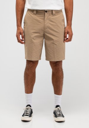 Mann trägt beige kniehohe Shorts, weiße Sportsocken, schwarze flache Turnschuhe und ein weißes T-Shirt, steht vor einem einfarbigen Hintergrund.