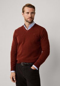 V-Ausschnitt Pullover aus tiefrotem Strickmaterial, mit gerippten Bündchen, getragen über einem hellblauen Kragenhemd. Kombiniert mit dunklen Hosen und einem schwarzen Gürtel.