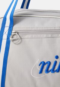 Mala desportiva cinza com alças listadas em azul e branco, bolso com fecho éclair e texto bordado em azul, apresentando um tecido texturizado.