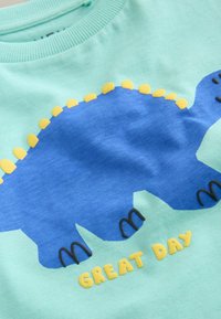 Türkisses puuvillane dressipluus sinise dinosauruse kujutisega, millel on kollased okkad, mustad detailid ja tekst "SUUREPÄRANE PÄEV" kollases.