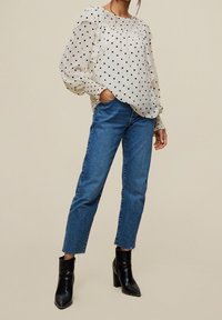 Durchsichtige weiße Bluse mit schwarzen Pünktchen, Rüschenärmel, kombiniert mit blauen Jeans und schwarzen Stiefeletten mit Absatz.