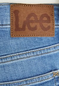 Világoskék farmeranyag, látható varrással, egy barna bőr folttal, amelyen a "Lee" logó egy laza betűtípussal látható.