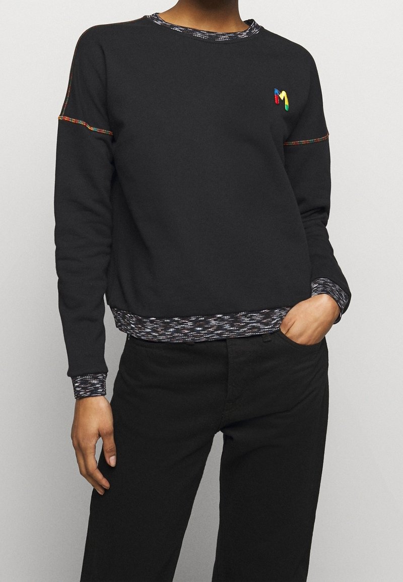 Persona vestida con una camiseta negra de manga larga con costuras multicolores y pantalones negros, con una mano en el bolsillo, recortada en el cuello y las caderas.