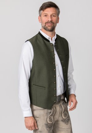 Mann trägt traditionelles bayerisches Outfit mit grüner Weste, weißem Hemd und beigen bestickten Lederhosen, steht vor einem einfarbigen Hintergrund.