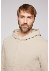 Sudadera con capucha beige con dos bolsillos en el pecho abotonados, hecha de tela suave. Diseño sencillo y casual con un ajuste relajado y capucha con cordón.