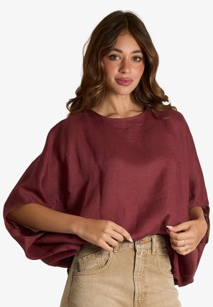 Donna con capelli castani ondulati che indossa una maglia bordeaux larga e pantaloni beige a vita alta, mentre sistema l'orlo della maglia.