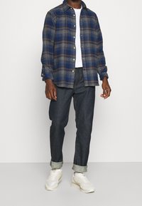 Blå rutigt flanellskjorta med knappar framtill, matchad med mörk denimjeans och vita sneakers. Flanellen har beige och marinblå detaljer.