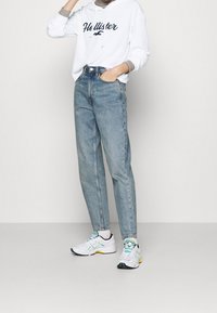 Hoodie blanc côtelé avec "Hollister" en bleu marine, jean taille haute bleu clair, et baskets blanches avec des accents verts et jaunes.