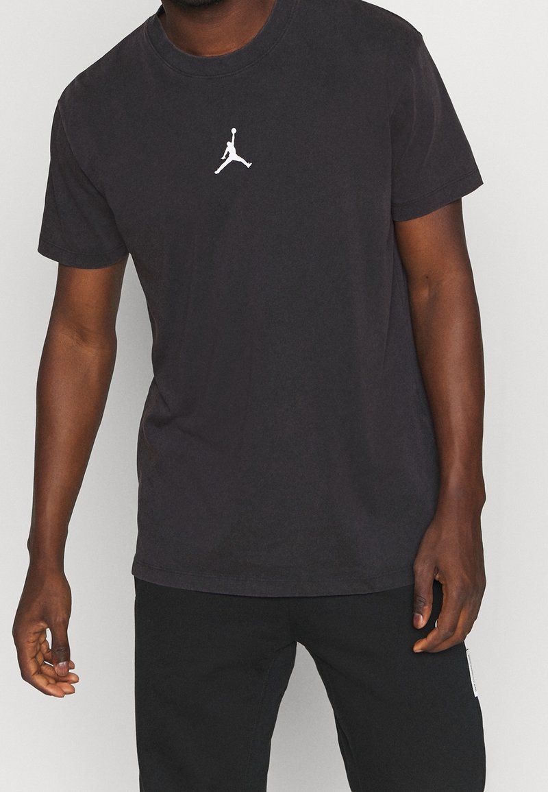 Man som bär en svart t-shirt med en liten vit Jumpman-logga på bröstet, i kombination med svarta byxor mot en enkel bakgrund.