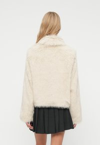 Žena s dlhými blond vlasmi, ktorá má na sebe smotanovo biely kabát z umelej kožušiny a tmavú plisovanú sukňu, zobrazená zozadu na bielom pozadí.