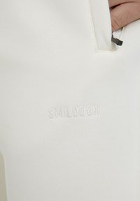 Weiße Baumwoll-Jogginghose mit glatter Textur, seitlicher Tasche mit Reißverschluss und geprägtem "SMILODOX"-Logo auf dem linken Oberschenkel.
