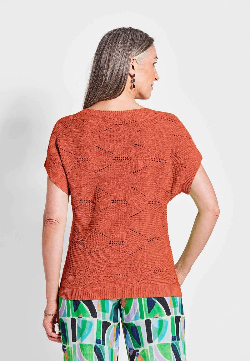 Rostfarbener kurzärmeliger Strickpullover mit einem geometrischen Lochmuster, kombiniert mit bunten gemusterten Hosen.