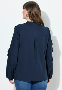 Marineblaue Bluse mit Rüschendetails an Ärmeln und Ausschnitt, strukturiertem Stoff und lockerer Passform. Der Rücken hat ein vertikales Nahtdetail.