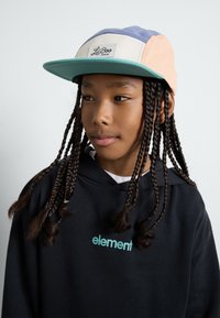 Mehrfarbige Kappe mit strukturiertem Design und einem Frontlabel, getragen mit einem schwarzen Kapuzenpullover mit Logo.