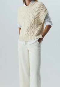 Krämfärgad kabelstickad tröja med rundad halsringning, bärs över en vit skjorta med krage, i kombination med ljusbeiga vida byxor.