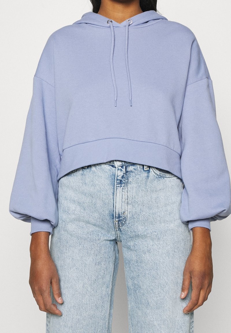 Personne portant un sweat à capuche court lavande avec des manches ballon et un jean taille haute délavé, debout devant un fond uni.