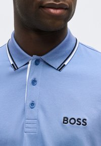 Άνδρας που φοράει ανοιχτό μπλε πόλο με γιακά με κουμπιά και το κέντημα "BOSS" στο στήθος, εμφανιζόμενος από το λαιμό μέχρι το μέσο του κορμού.