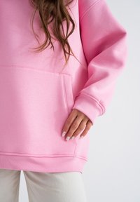 Felpa rosa con tasca frontale, polsini e orlo a coste. Il tessuto appare morbido con una superficie liscia. Mano appoggiata sulla tasca.