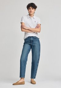 Chemise polo blanche à manches courtes avec un logo noir, associée à un jean en denim bleu ample et des chaussures plates couleur tan. Design simple, look décontracté.