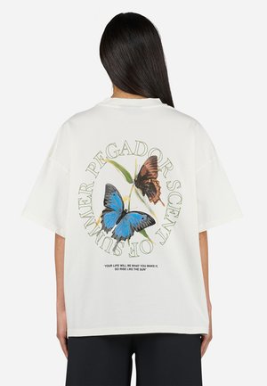 Hvit bomull t-skjorte med et stort sommerfugl grafikk i blått og brunt bak, med grønn tekst som står "PEGADOR SCENT OF SUMMER."
