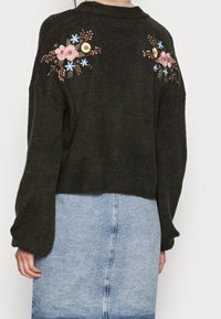 Maglione nero con ricami floreali colorati sulle spalle, indossato con una gonna di jeans azzurra a vita alta, visto da dietro.