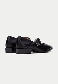 Zapatos de cuero negros con un tacón bajo y cuadrado, punta redondeada y una correa decorativa con hebilla a lo largo del empeine. Textura suave y diseño clásico.