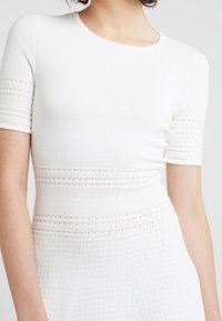 Haut tricoté blanc à manches courtes, présentant un motif texturé et des rayures horizontales avec de petites découpes. Encolure ronde et coupe ajustée.