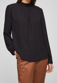 Schwarze Bluse mit langen Ärmeln und hohem Ausschnitt, gefälteltem Detail an der Vorderseite und strukturierten Manschetten, kombiniert mit braunen Satin-Hosen.