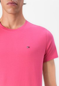 Homme portant un t-shirt à manches courtes rose vif avec un petit logo rectangulaire sur la poitrine gauche, sur fond blanc.