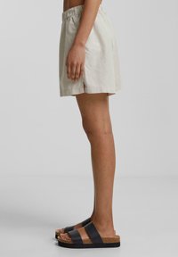 Shorts en lin beige clair avec une taille élastique, des fentes latérales et une coupe ample, associés à des sandales à glissière noires et beiges.
